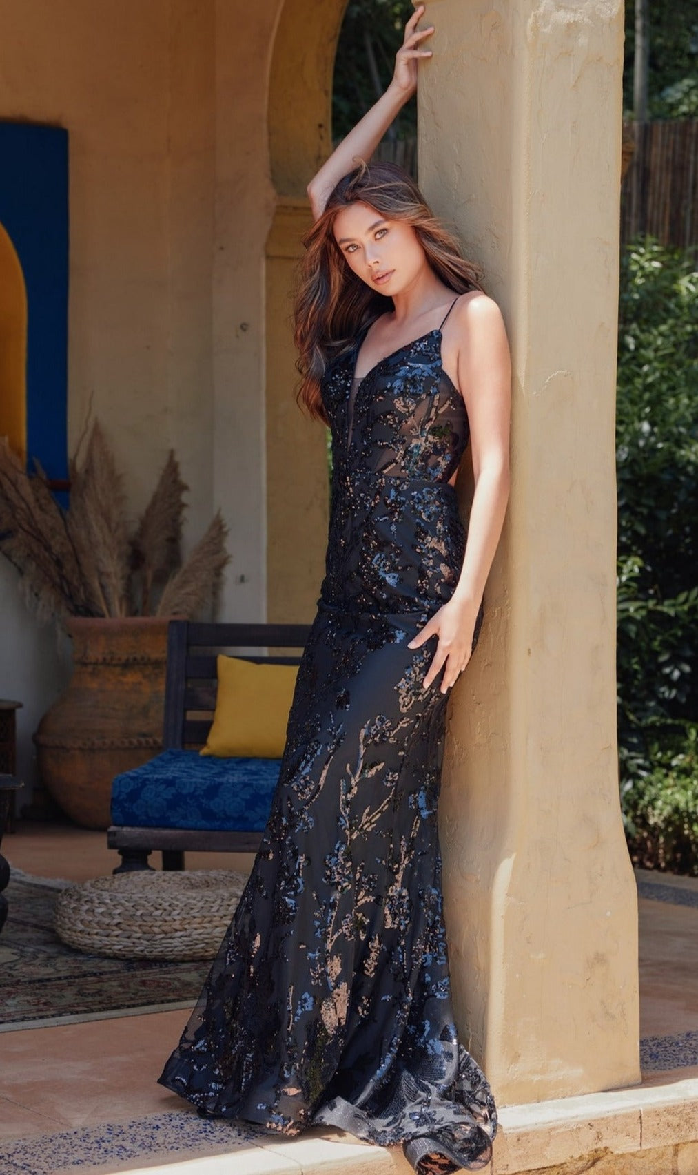 A woman in the Strappy-Back Long Sequin-Print Prom Dress 2411 leans against a pillar outdoors, surrounded by rustic decor and lush greenery.